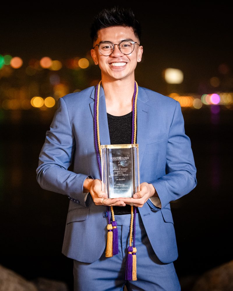 Les Balibat in a blue suit and graduation cords, holding his MBA Student of the Year award at night by the water